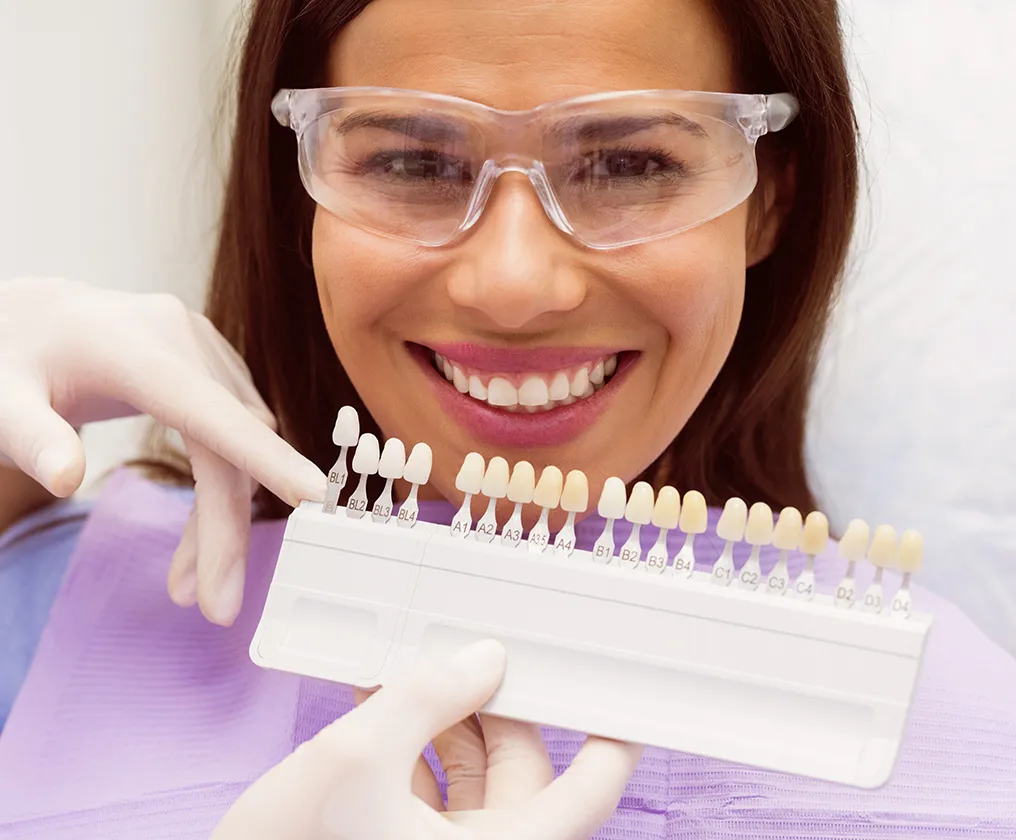 A smiling patient in protective glasses having her tooth shade matched against a whitening shade guide