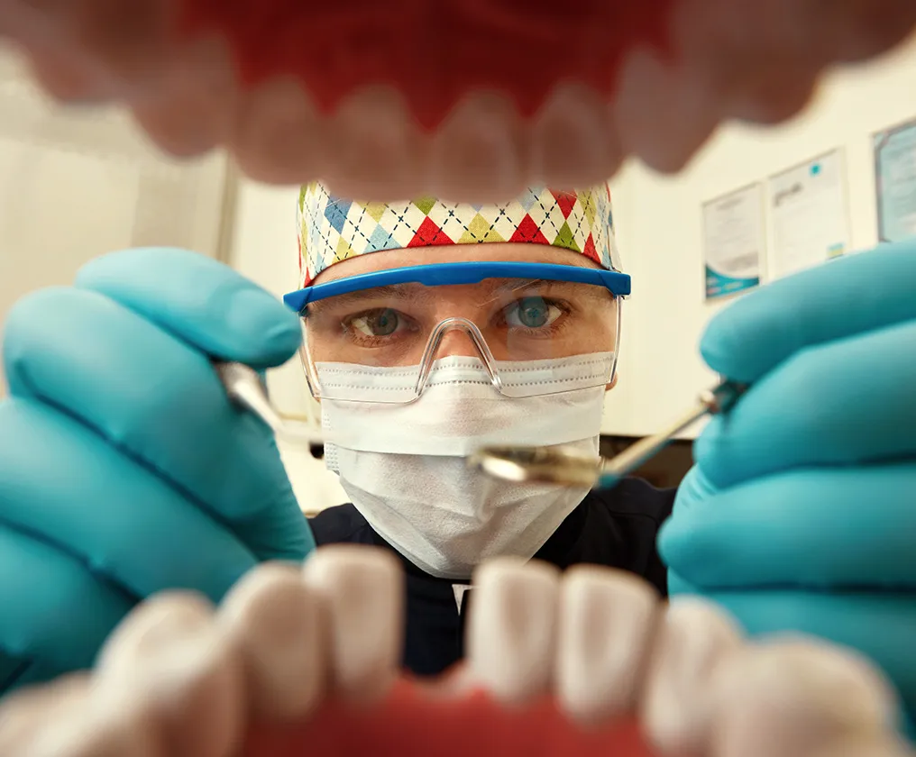 A dentist in a mask and protective glasses holding a mirror and probe, seen from the patient's point of view