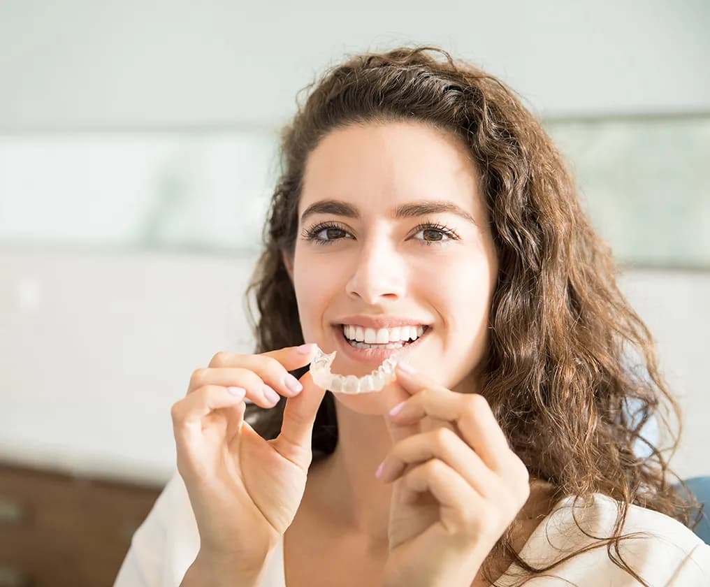 A smiling woman holding a clear aligner up in front of her teeth